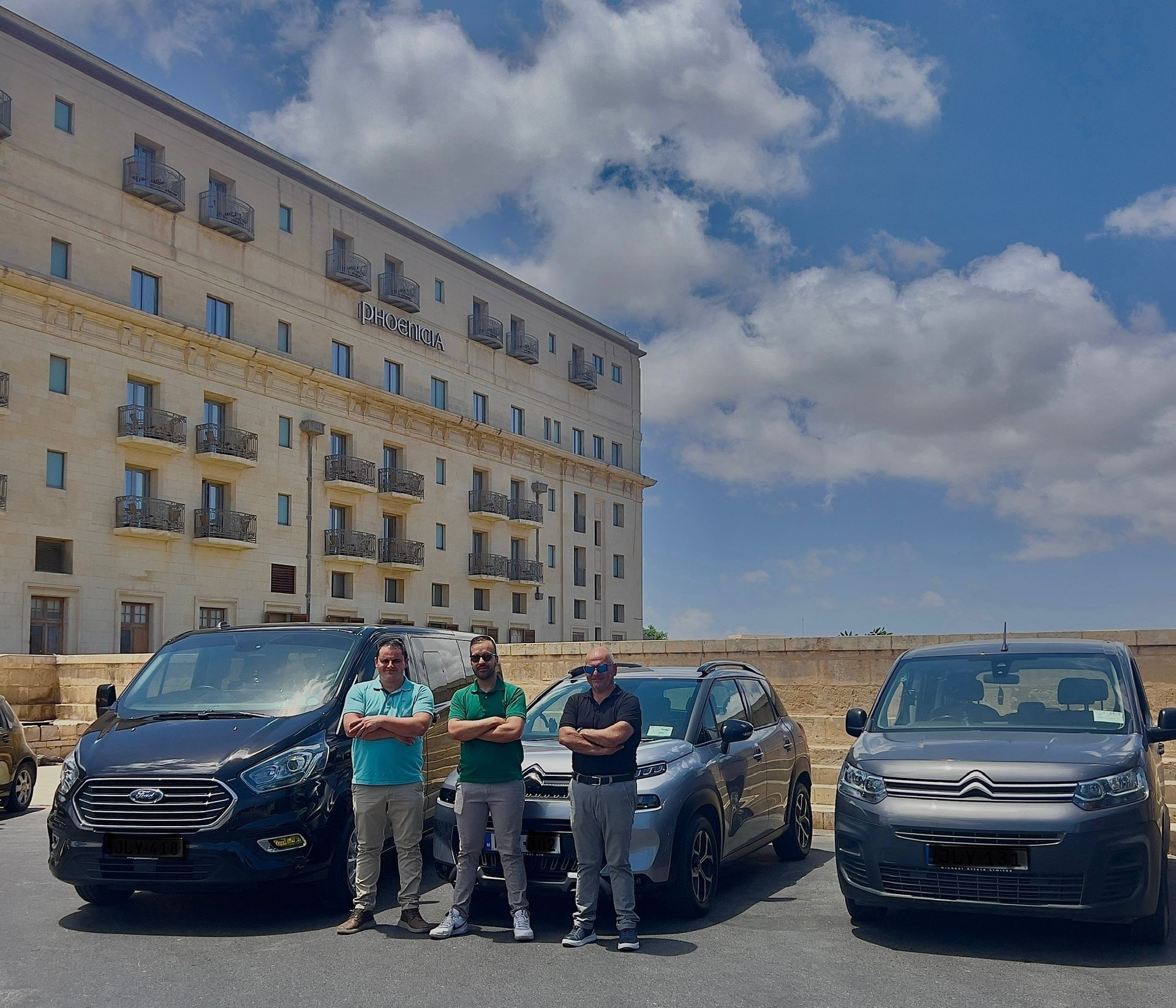 Three men standing with arms crossed in front of parked cars near a large building on a sunny day.
