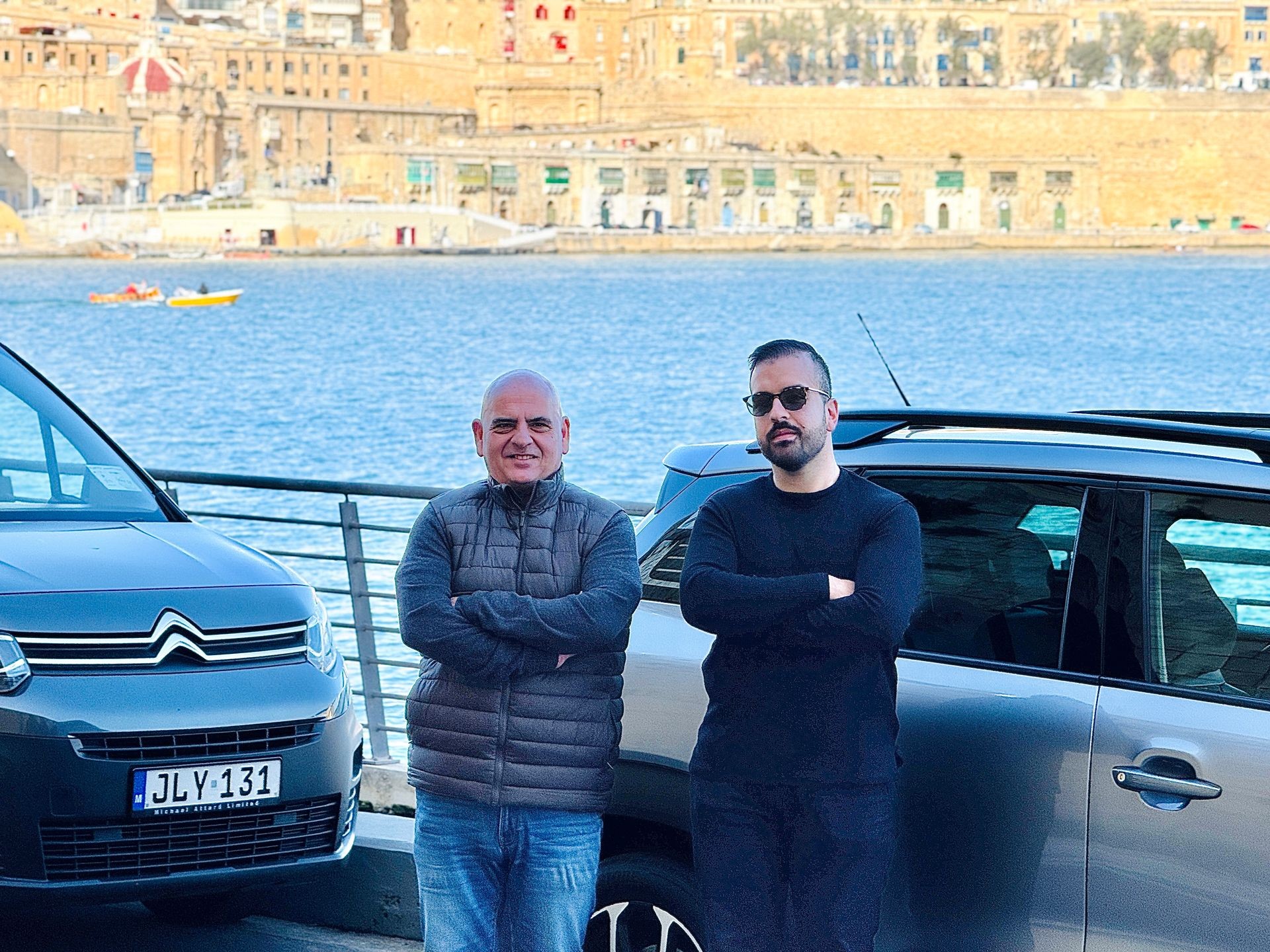 Two men standing in front of parked cars by a waterfront with a historic building in the background.