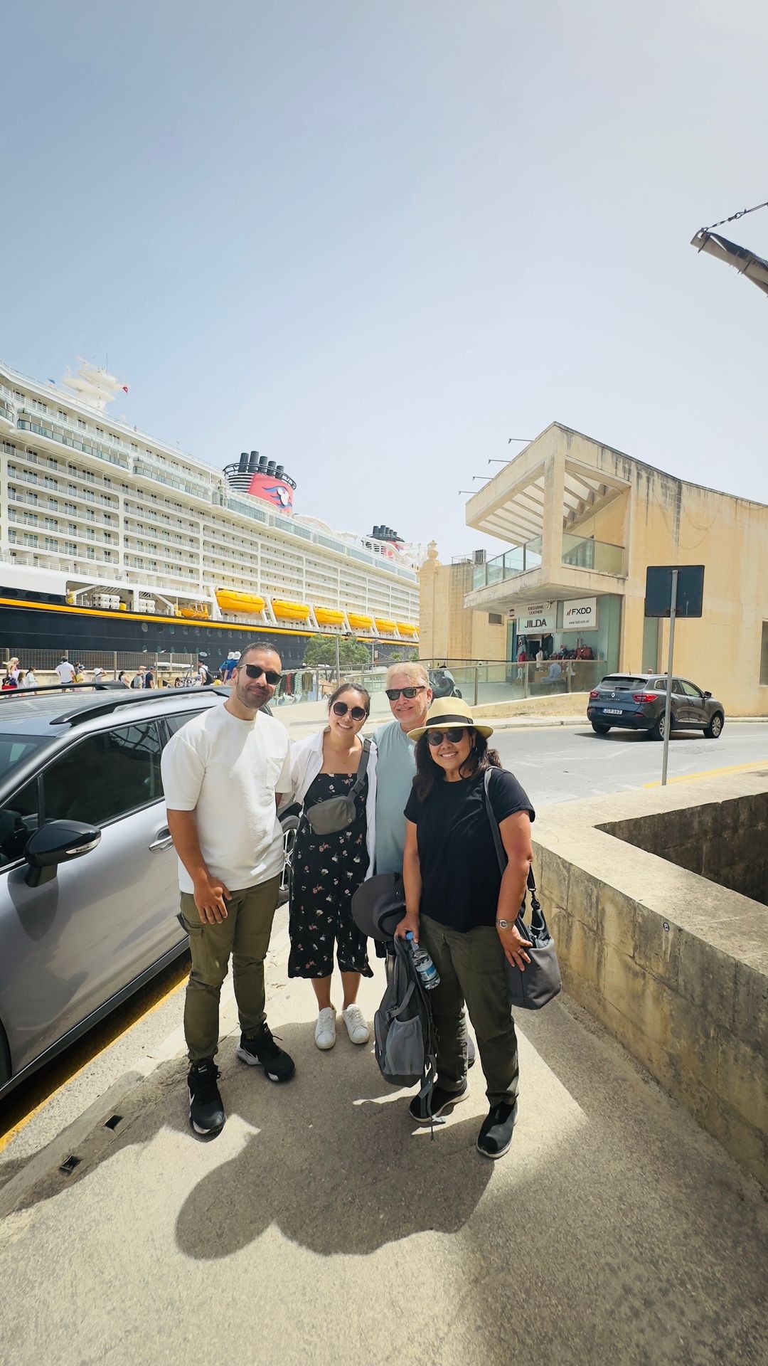 Group of four people on a sunny street with a large cruise ship and buildings in the background.